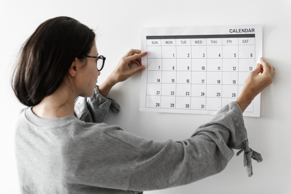 A imagem contém uma mulher colando um calendário referente a um mês em uma parede para dar início ao seu planejamento de gestão do tempo e produtividade.
