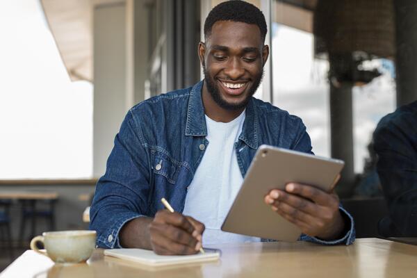 A imagem contém um empresário sorridente fazendo notas sobre o plano de gerenciamento de riscos de sua startup, enquanto segura um tablet com a outra mão.